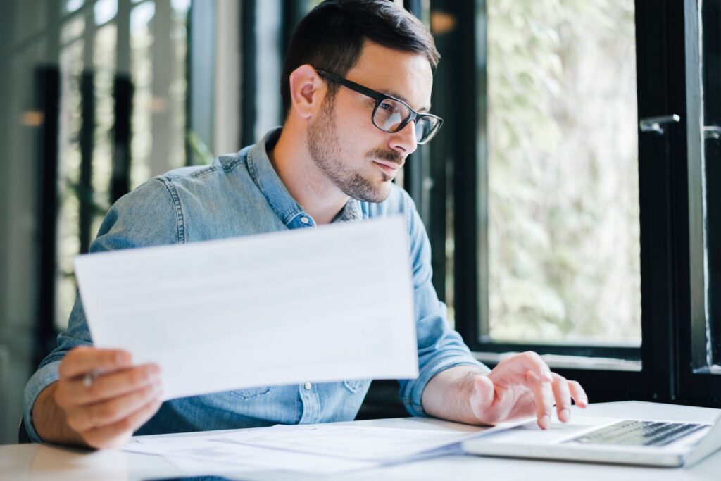 A man wearing glasses reviews a document while working on a laptop at a desk in a bright office
