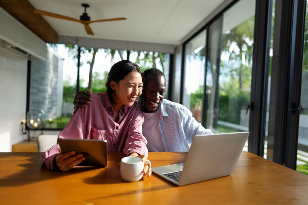 Two people smiling while looking at a laptop, with one holding a tablet and a coffee mug on the table