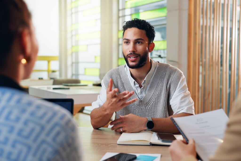 Person speaking and gesturing during a meeting with two people seated across a table