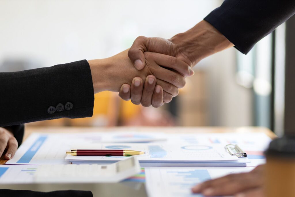 Two business professionals shake hands across a desk with financial reports, charts, and a pen, symbolizing a successful business agreement