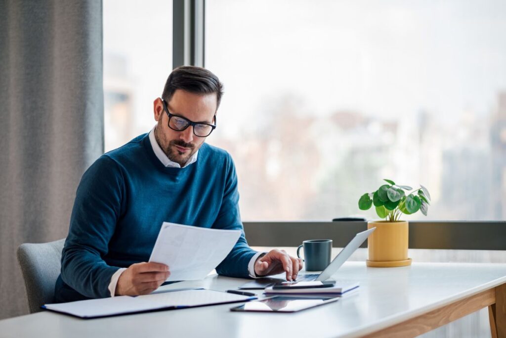 Business professional reviewing financial documents while working on a laptop at a desk in a modern office