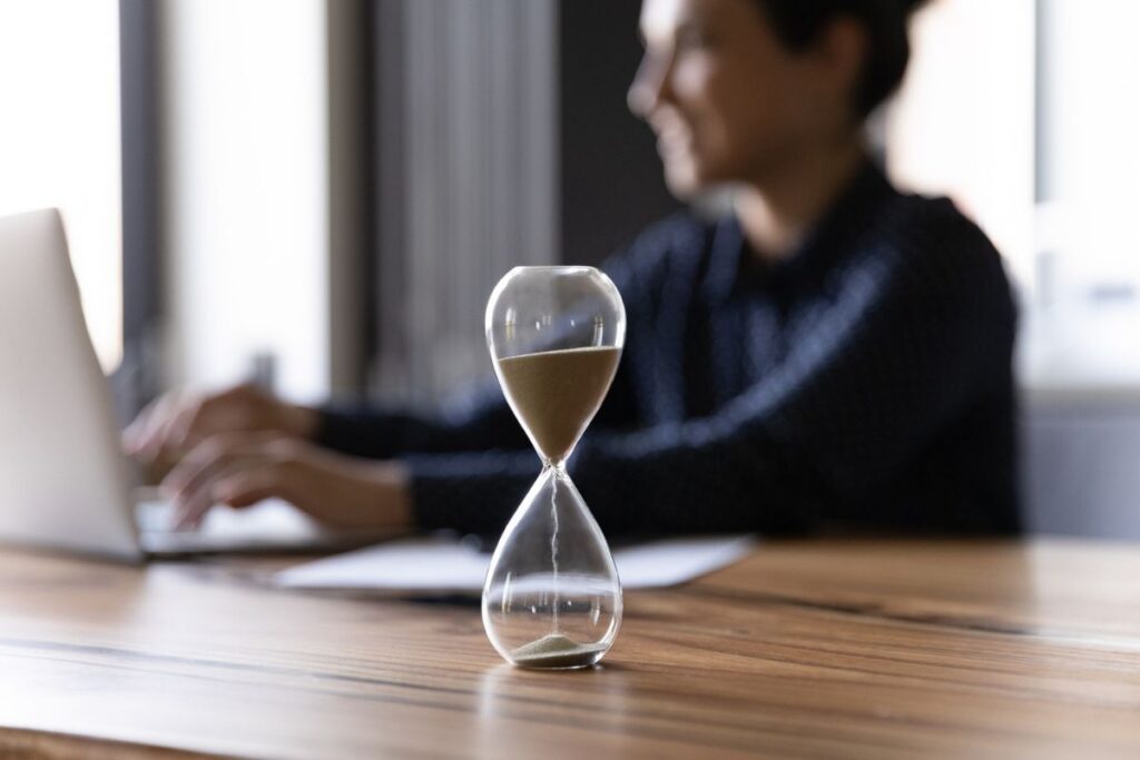 Hourglass on a desk in the foreground while a business professional works on a laptop in the background