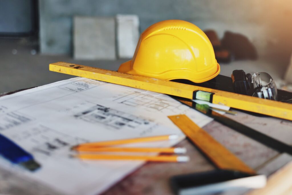 Construction plans, measuring tools, and a yellow hard hat on a worktable.